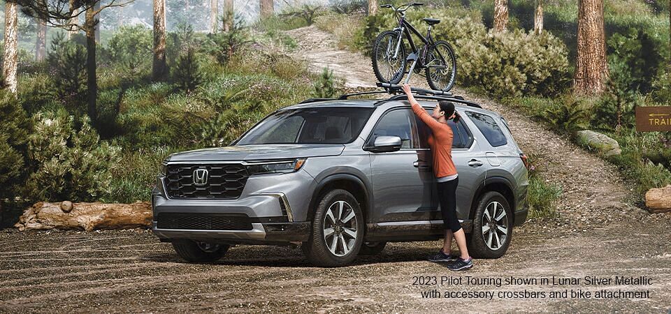 Woman placing bicycle on 2023 Honda Pilot shown in Lunar Silver metallic with accessory crossbars and bike attachment standing on a forest parking.