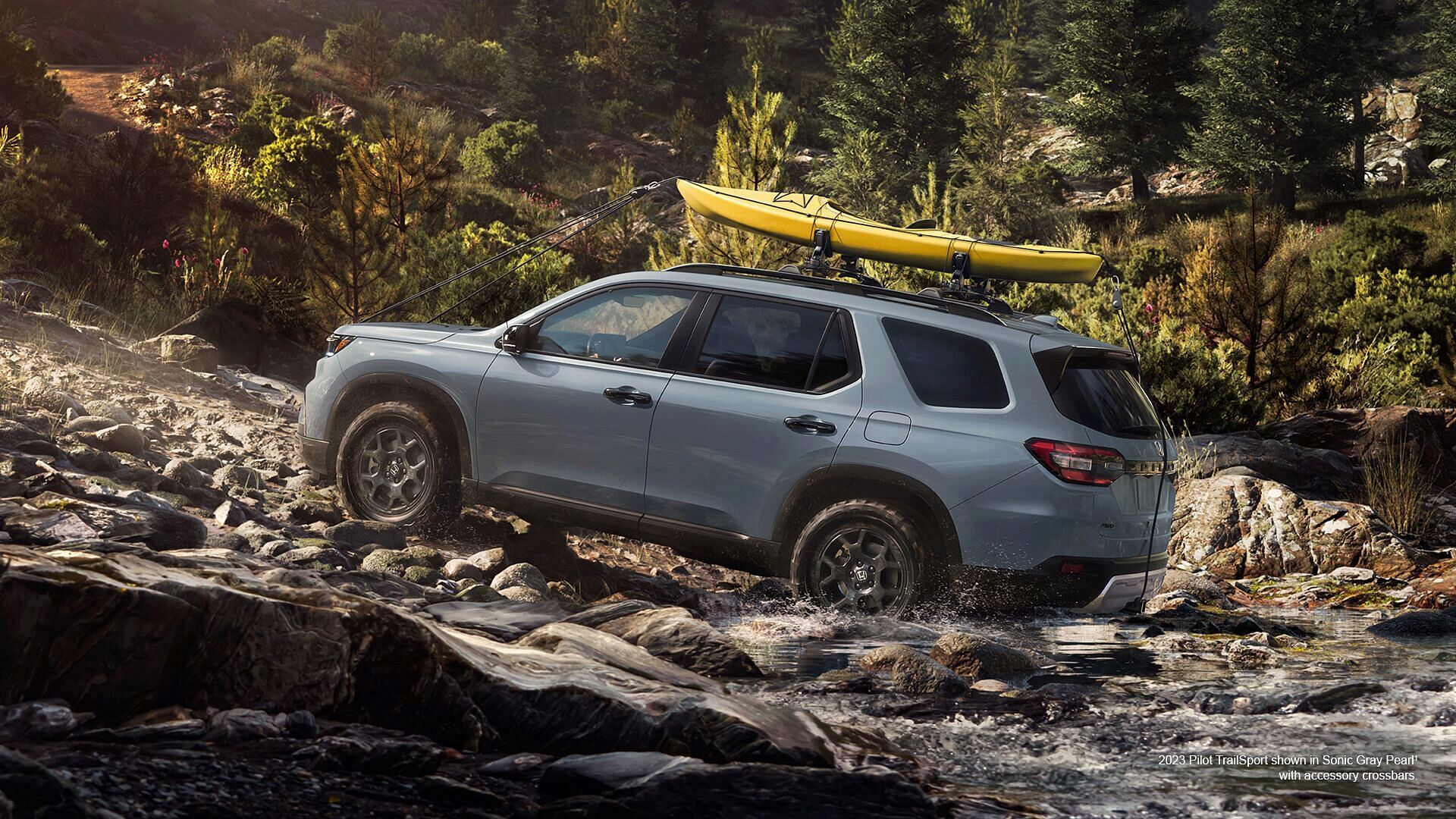 A Sonic Gray Pearl Honda Pilot TrailSport 2023 with accessory crossbars and kayak attachment driving through forest stream